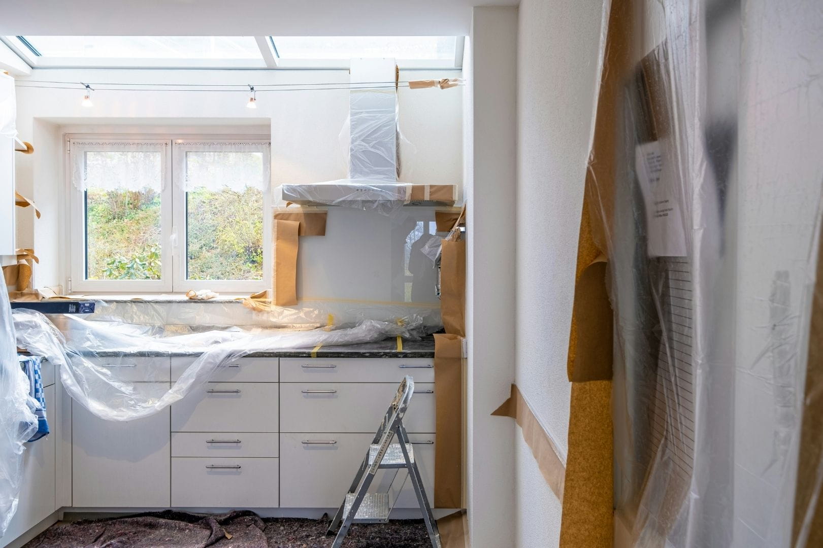 Cuisine en cours de rénovation avec protections plastiques sur les meubles blancs et matériel de peinture.