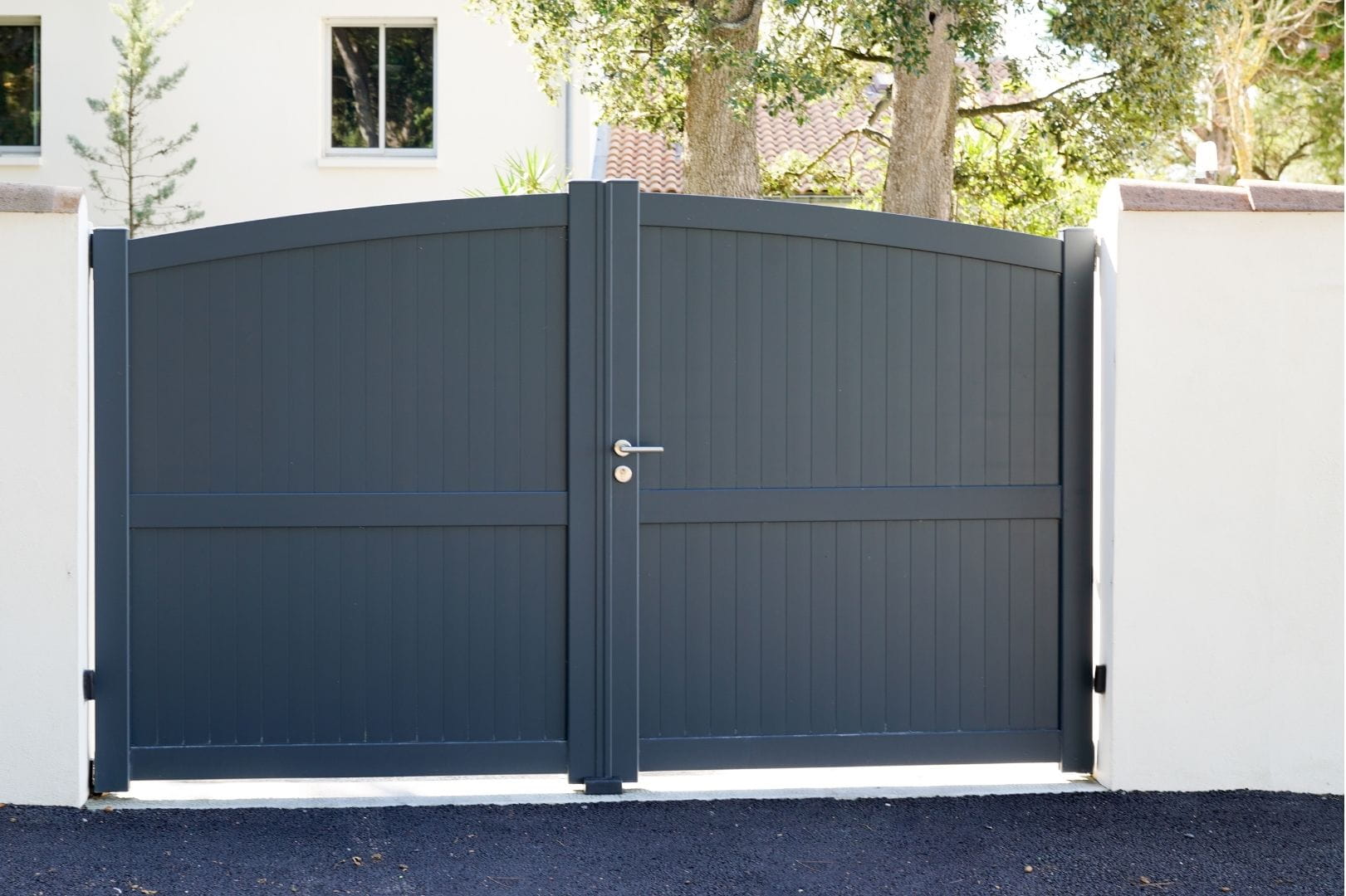 Portail battant moderne en aluminium gris posé entre deux piliers maçonnés blancs à l'entrée d'une maison.